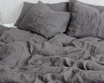 May include: A bed dressed with grey linen bedding, including a duvet cover and pillows. The fabric has a wrinkled texture, and the pillows are arranged on top of the bed. The scene is lit with natural light, creating soft shadows.