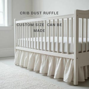 Könnte beinhalten: Ein weißer Bettrüschen-Volant für ein Kinderbett, befestigt an einem weißen Gitterbett. Der Text CRIB DUST RUFFLE und CUSTOM SIZE CAN BE MADE ist in schwarzer Schrift sichtbar. Der Volant hat ein gerafftes Stoffdesign am Bettgestell.