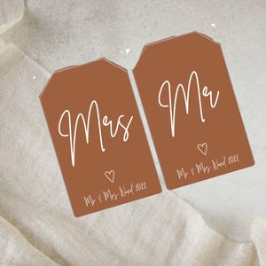 Puede incluir: Dos etiquetas de regalo marrones con escritura blanca que dice "Mrs." y "Mr." con un pequeño corazón y el texto "Mr. & Mrs. Wood 2022" debajo.