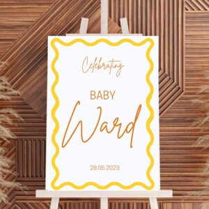 Puede incluir: Un letrero blanco sobre un caballete con un borde amarillo ondulado. El letrero dice "Celebrating BABY Ward" en una fuente manuscrita, con la fecha "28.05.2023" en la parte inferior. El fondo es una pared de madera con plantas secas a ambos lados.