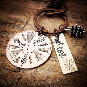 May include: A copper keychain with a circular charm featuring a design of ferns and the text "get lost". The keychain also includes a small pine cone charm and a brass tag with the text "get lost" and a compass design.
