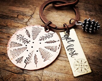 May include: A copper keychain with a circular charm featuring a design of ferns and the text "get lost". The keychain also includes a small pine cone charm and a brass tag with the text "get lost" and a compass design.