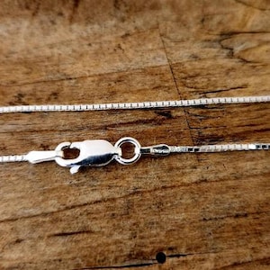 May include: A silver chain necklace with a lobster clasp closure. The chain is made of small, square links.