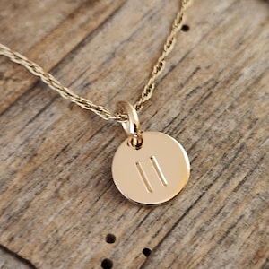 May include: A gold-toned necklace featuring a small, circular pendant. The pendant is engraved with two vertical parallel lines. The necklace is attached to a delicate, twisted gold-toned chain that connects to the top of the circular charm.