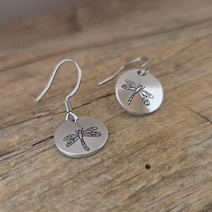 May include: A pair of silver-colored dragonfly earrings. Each earring features a circular pendant with a detailed black dragonfly design. The earrings have a simple hook closure and are displayed on a wooden surface.