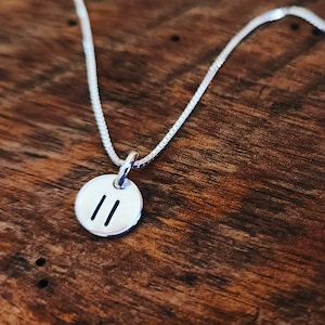 May include: A silver necklace with a round pendant featuring the double bar symbol. The necklace is on a wooden surface.