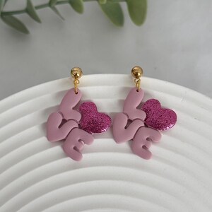 May include: Dangle earrings with a romantic design. Each earring spells "LOVE" in a soft pink, with a glittery magenta heart. The earrings have gold-coloured ball studs.