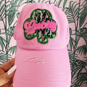 以下が含まれることがあります： ピンク色のダメージ加工が施されたベースボールキャップ。スパンコールで作られたワッペンはクローバーの形で、ピンクのスパンコールで「groovy」の文字が描かれています。キャップにはダメージ加工が施され、つばはカーブしています。