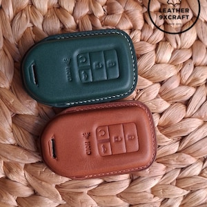 May include: Two leather key fob covers, one forest green and one brown, with white stitching. Each cover has embossed buttons and the word "HOLD". The covers are on a woven background. The top right corner has a logo that says "LEATHER 9XCRAFT".
