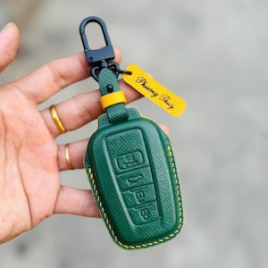 Op de afbeelding: Groene leren autosleutelhoes met gele stiksels en een zwarte metalen sluiting. De sleutelhanger heeft reliëfknoppen en een geel label met de tekst "Phuong Thuy".