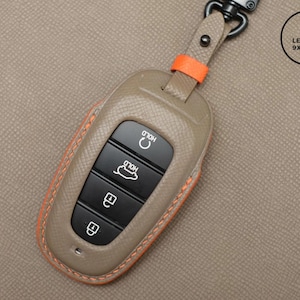 May include: A tan leather key fob cover with orange stitching and accents. The key fob has black buttons with white icons and the word "HOLD". A metal clasp and a small orange strap are attached to the top. The Leather 9xCraft logo is in the upper right corner.