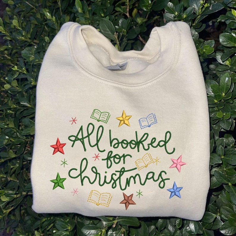 May include: A cream-colored crewneck sweatshirt featuring green embroidered text that reads All booked for Christmas. The design is accented with small, colorful embroidered stars and open book icons in red, yellow, blue, and green hues.