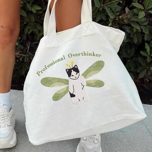 May include: White canvas tote bag with the text "Professional Overthinker" and an embroidered image of a black and white cat with green wings.