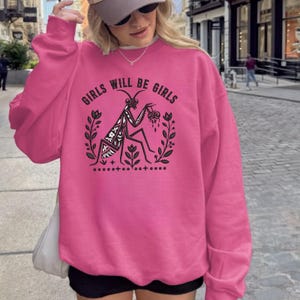 May include: A bright pink long-sleeve sweatshirt featuring a black graphic print of a praying mantis surrounded by floral sprigs. The text "GIRLS WILL BE GIRLS" is arched above the insect, which is holding a small heart-shaped object.