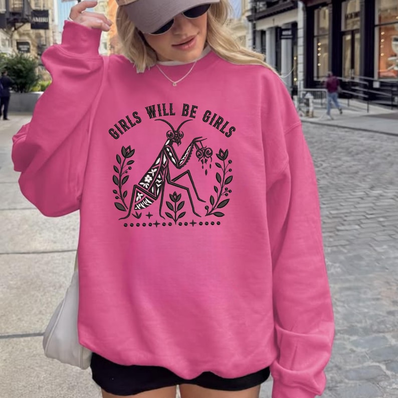 May include: A bright pink long-sleeve sweatshirt featuring a black graphic print of a praying mantis surrounded by floral sprigs. The text "GIRLS WILL BE GIRLS" is arched above the insect, which is holding a small heart-shaped object.