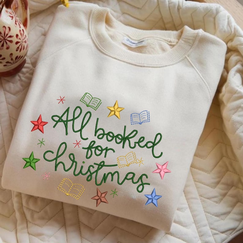 May include: A cream-colored crewneck sweatshirt featuring green embroidered text that reads All booked for Christmas. The design is accented with colorful embroidered stars and small open book icons in red, yellow, blue, pink, and green thread.
