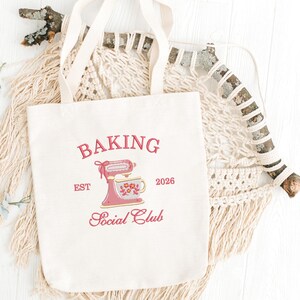 Op de afbeelding: Cr&egrave;mekleurige draagtas met de woorden "BAKING Social Club" in roze script. Een roze keukenmixer en bloementheekopje staan in het midden. De tas heeft twee cr&egrave;mekleurige handvatten en wordt gepresenteerd op een witte ondergrond.