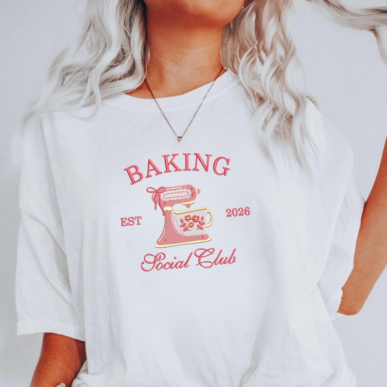 Op de afbeelding: Wit T-shirt met een roze afbeelding van een keukenmixer en een bloemenmok. De tekst luidt "BAKING Social Club EST 2026" in een roze, vintage-ge&iuml;nspireerd lettertype. Het shirt heeft korte mouwen.