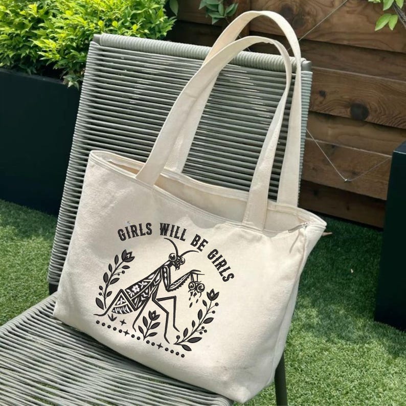May include: A cream-colored canvas tote bag featuring a black graphic print of a praying mantis surrounded by floral elements. The text "GIRLS WILL BE GIRLS" is printed in a curved arch above the insect illustration on the front of the bag.