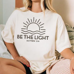 K&ouml;nnte beinhalten: Cremefarbenes T-Shirt mit dem Text "BE THE LIGHT" und dem Bibelvers Matth&auml;us 5:14. Das Design zeigt eine Sonne und Wellen in einem minimalistischen Stil. Das Shirt hat eine lockere Passform.