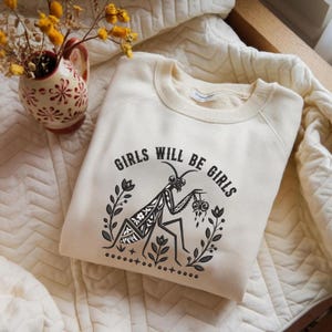 May include: A cream-colored crewneck sweatshirt featuring a black graphic print of a praying mantis surrounded by floral elements and the text "GIRLS WILL BE GIRLS" arched above the insect. The design includes small star and dot accents.