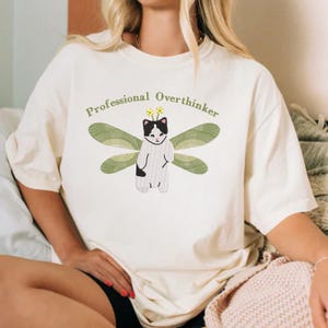May include: White t-shirt with a graphic of a cat with wings and flowers on its head. The text "Professional Overthinker" is printed above the cat.
