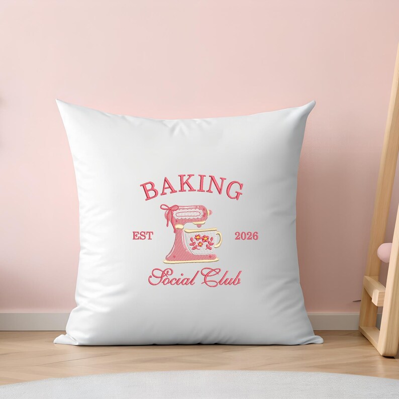 Op de afbeelding: Wit kussen met een roze en rood ontwerp. Het ontwerp bevat de tekst "BAKING Social Club EST 2026" boven een illustratie van een roze keukenmixer en een bloemenkopje.
