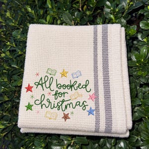May include: A cream-colored waffle-weave kitchen towel featuring two vertical gray stripes and embroidered text that reads All booked for Christmas in green, surrounded by small colorful stars and open book icons in various colors.