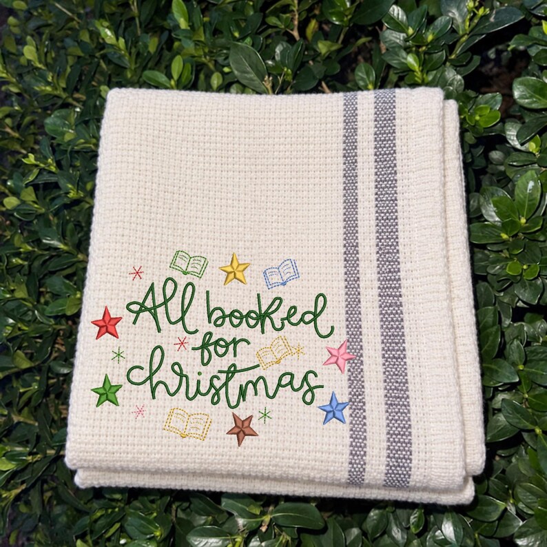 May include: A cream-colored waffle-weave kitchen towel featuring two vertical gray stripes and embroidered text that reads All booked for Christmas in green, surrounded by small colorful stars and open book icons in various colors.