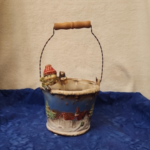 May include: A ceramic bucket-shaped planter with a winter scene design. The bucket has a blue base with a painted village scene and a figure wearing a red hat. It has a wooden handle and wire supports.