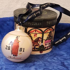May include: Cream-colored Christmas ornament with a figure in a red coat, the numbers "20 01", and the words "Merry Christmas". It is next to a round box with a black lid and a ribbon with the words "Ralph Lauren".