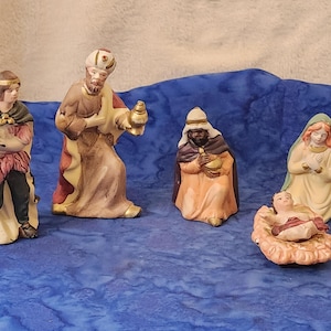 May include: A ceramic nativity scene featuring five figures. The figures include the three wise men, Mary, and baby Jesus in a manger. The figures are painted in various colors, including brown, beige, and gold. The scene is set against a blue background.