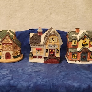 May include: Three ceramic Christmas village buildings: a tavern, a bakery, and a pub. The tavern has a green roof and the word "Tavern". The bakery is yellow with a red awning and the word "Bakery". The pub is red with the words "Beer Pub".