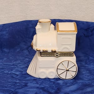 May include: A white ceramic train figurine with gold accents. The train features a detailed design, including wheels, a smokestack, and a decorative flower. The figurine is set against a blue background.