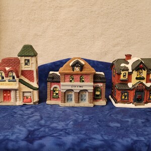 May include: Three ceramic Christmas village buildings: a toy shop, a train station, and a pub. The toy shop is red and white with a green roof. The train station is brick-colored with a clock. The pub is red with snow.