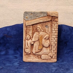 May include: A ceramic relief sculpture depicting a scene of the nativity. The sculpture features a woman and a child standing in front of a doorway, with a brick wall and a star-filled sky above.