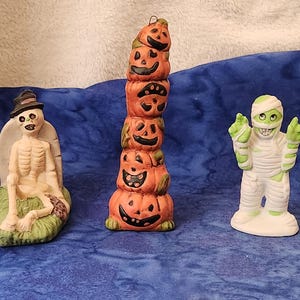 May include: Three Halloween-themed figurines: a skeleton in a hat sitting by a tombstone, a stack of orange jack-o'-lanterns, and a white mummy with green accents. The figurines are on a blue surface.