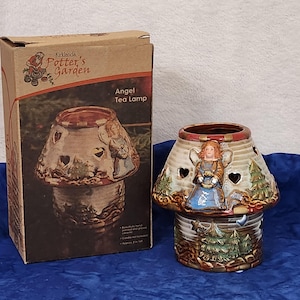 May include: A ceramic tea light holder shaped like a house with an angel design. The holder is decorated with a brown, green, and white glaze. The box reads "Kirkland's Potter's Garden Angel Tea Lamp".