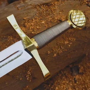 Pode incluir: Uma espada de estilo medieval com uma lâmina de aço polido e uma guarda dourada. O cabo é envolto em um material escuro e texturizado. O pomo e a guarda são dourados. A espada repousa sobre uma superfície de madeira.