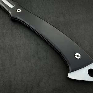 Peut inclure: Gros plan sur un manche de couteau noir avec une lame argent&eacute;e. La poign&eacute;e a un design incurv&eacute; avec une finition brillante et une extr&eacute;mit&eacute; argent&eacute;e avec un trou circulaire. Le couteau est pos&eacute; sur un fond sombre.