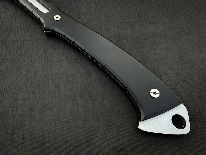 Peut inclure: Gros plan sur un manche de couteau noir avec une lame argent&eacute;e. La poign&eacute;e a un design incurv&eacute; avec une finition brillante et une extr&eacute;mit&eacute; argent&eacute;e avec un trou circulaire. Le couteau est pos&eacute; sur un fond sombre.