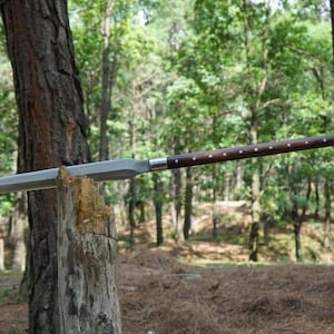 K&ouml;nnte beinhalten: Ein Speer mit einem langen, polierten Holzschaft und einer silbernen Klinge. Der Schaft ist dunkelbraun mit kleinen, gleichm&auml;&szlig;ig verteilten silbernen Akzenten. Der Speer ruht auf einem verwitterten Holzpfosten in einer Waldumgebung.