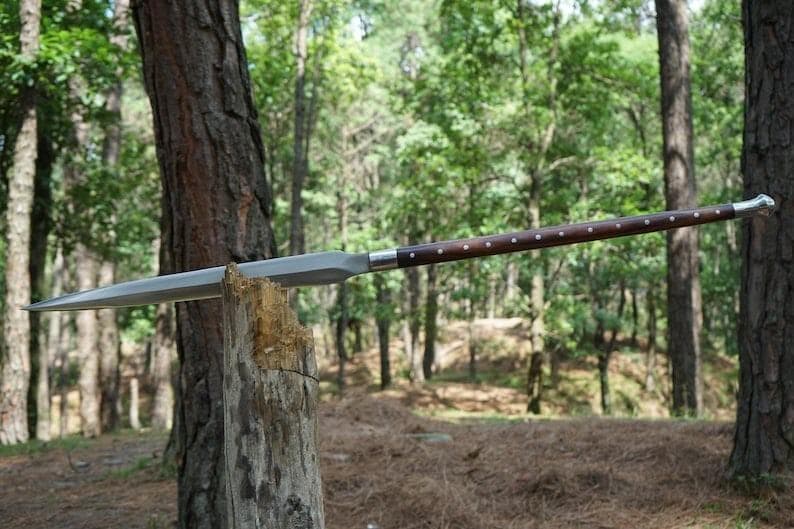 K&ouml;nnte beinhalten: Ein Speer mit einem langen, polierten Holzschaft und einer silbernen Klinge. Der Schaft ist dunkelbraun mit kleinen, gleichm&auml;&szlig;ig verteilten silbernen Akzenten. Der Speer ruht auf einem verwitterten Holzpfosten in einer Waldumgebung.