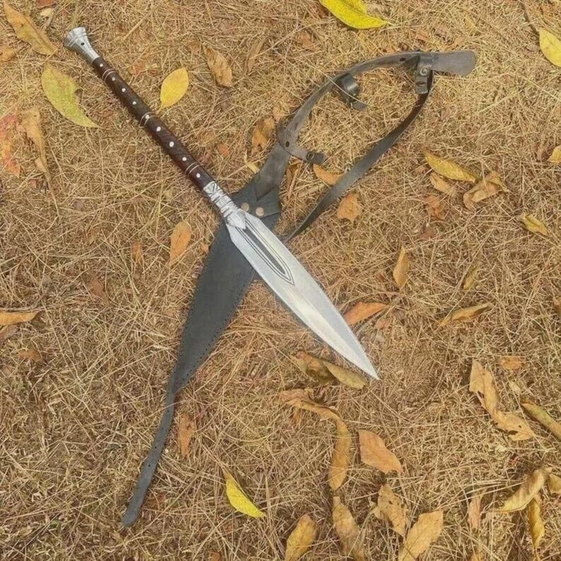 May include: A spear with a silver blade and a brown wooden handle with silver accents. The spear is resting on a bed of dry grass and leaves, with a black leather sheath and a metal harness.