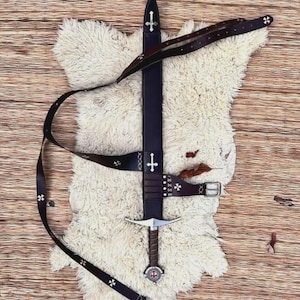May include: A dark brown leather sword scabbard and matching belt featuring silver-toned cross emblems. The scabbard has a pointed tip and a metal cross inlay, while the belt includes a metal buckle and small cross-shaped decorative studs.