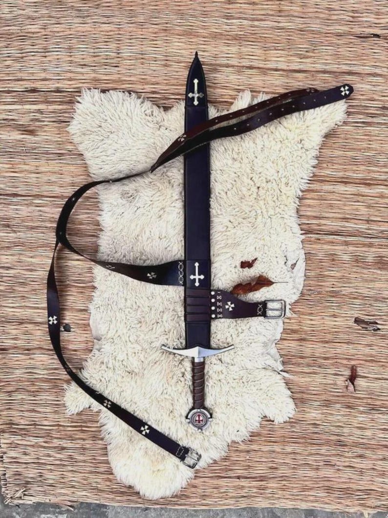 May include: A dark brown leather sword scabbard and matching belt featuring silver-toned cross emblems. The scabbard has a pointed tip and a metal cross inlay, while the belt includes a metal buckle and small cross-shaped decorative studs.
