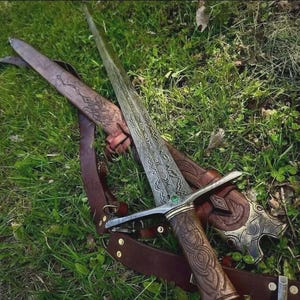 Pode incluir: Uma espada ornamentada com uma l&acirc;mina e punho detalhados, repousando sobre uma bainha e correia de couro. A espada apresenta padr&otilde;es intrincados e uma guarda prateada. A bainha &eacute; marrom com desenhos esculpidos, na grama verde.