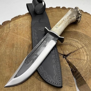 Puede incluir: Un cuchillo de caza con una hoja de acero pulido y un mango de asta. El cuchillo descansa sobre una superficie de madera, con una funda de cuero negro. La funda tiene patrones en relieve y una trabilla para el cinturón. La hoja mide unos 25 cm.