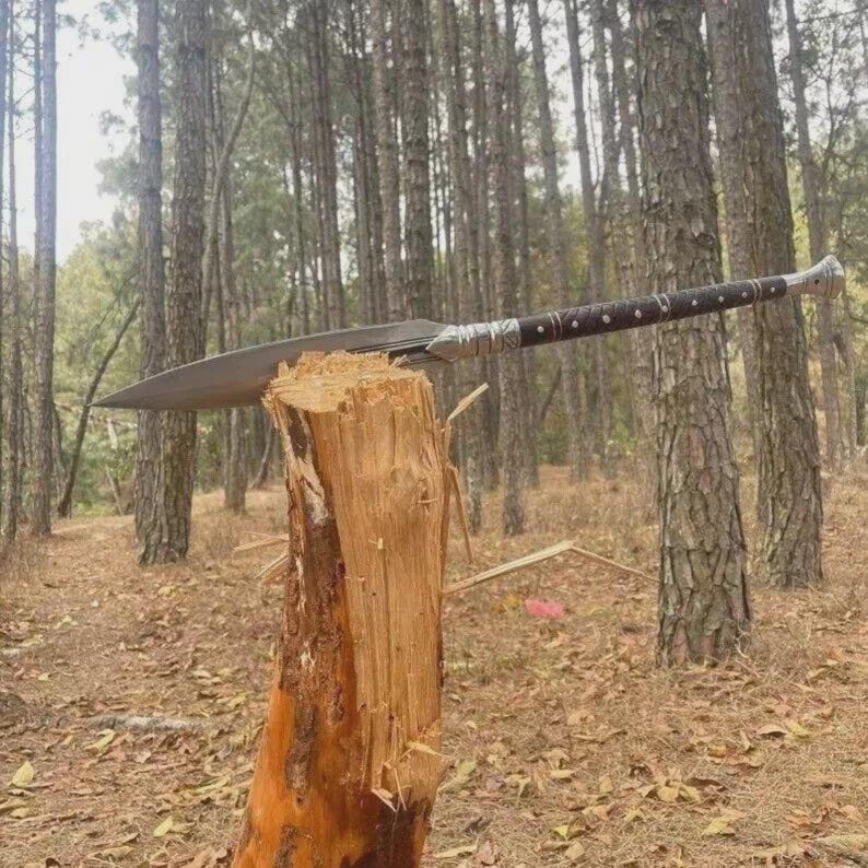 May include: A large, bladed spear with a dark wooden handle and silver accents, resting on a tree stump in a forest setting. The spearhead is wide and sharp, and the handle is adorned with small, decorative rivets. The background features tall trees and fallen leaves.