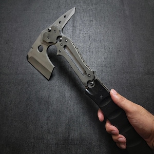 Puede incluir: Un tomahawk t&aacute;ctico con una hoja gris y un mango negro. El tomahawk presenta una cabeza de hacha afilada, una punta y un agarre texturizado para un manejo seguro. El dise&ntilde;o incluye m&uacute;ltiples tornillos y un agujero cerca de la cabeza del hacha.